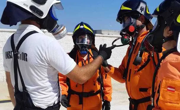 Bomberos/Alumnos vestidos de naranja, con cascos oscuros y mascaras puestas con un monitor con camiseta y cascos blancos