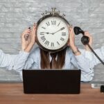 Mujer de pelo largo con las manos levantadas y en la cabeza un reloj sentada en una mesa con un ordenador delante