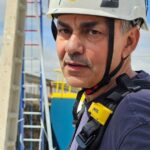 El profesor de la academia con casco blanco y camiseta azul en los exteriores de un almacén haciendo un curso de trabajos en altura.