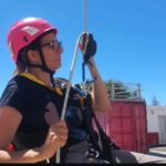 La periodista Lorena Díaz haciendo un curso de trabajos en altura con arnés y casco rojos en un sitio exterior.