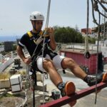 Alumno con casco blanco, camiseta oscura, y pantalón blanco el que lleva botas de seguridad. Está colgado de un arnés con todas las cuerdas de seguridad sobre una barandilla.