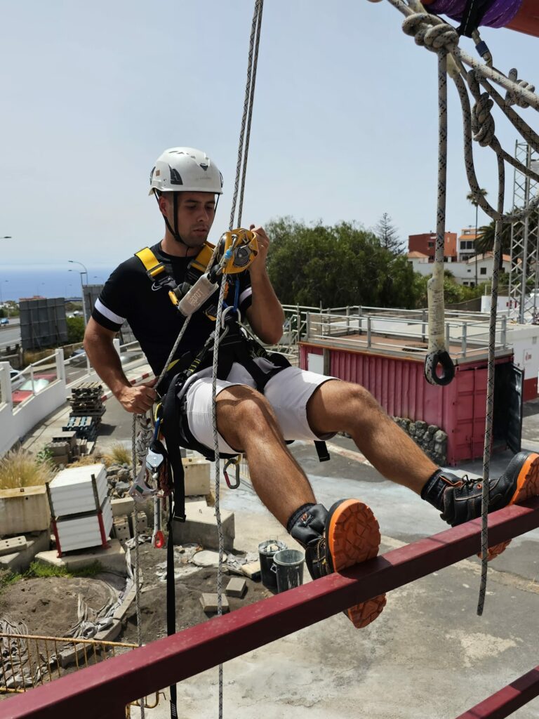 Alumno con casco blanco, camiseta oscura, y pantalón blanco el que lleva botas de seguridad. Está colgado de un arnés con todas las cuerdas de seguridad sobre una barandilla.